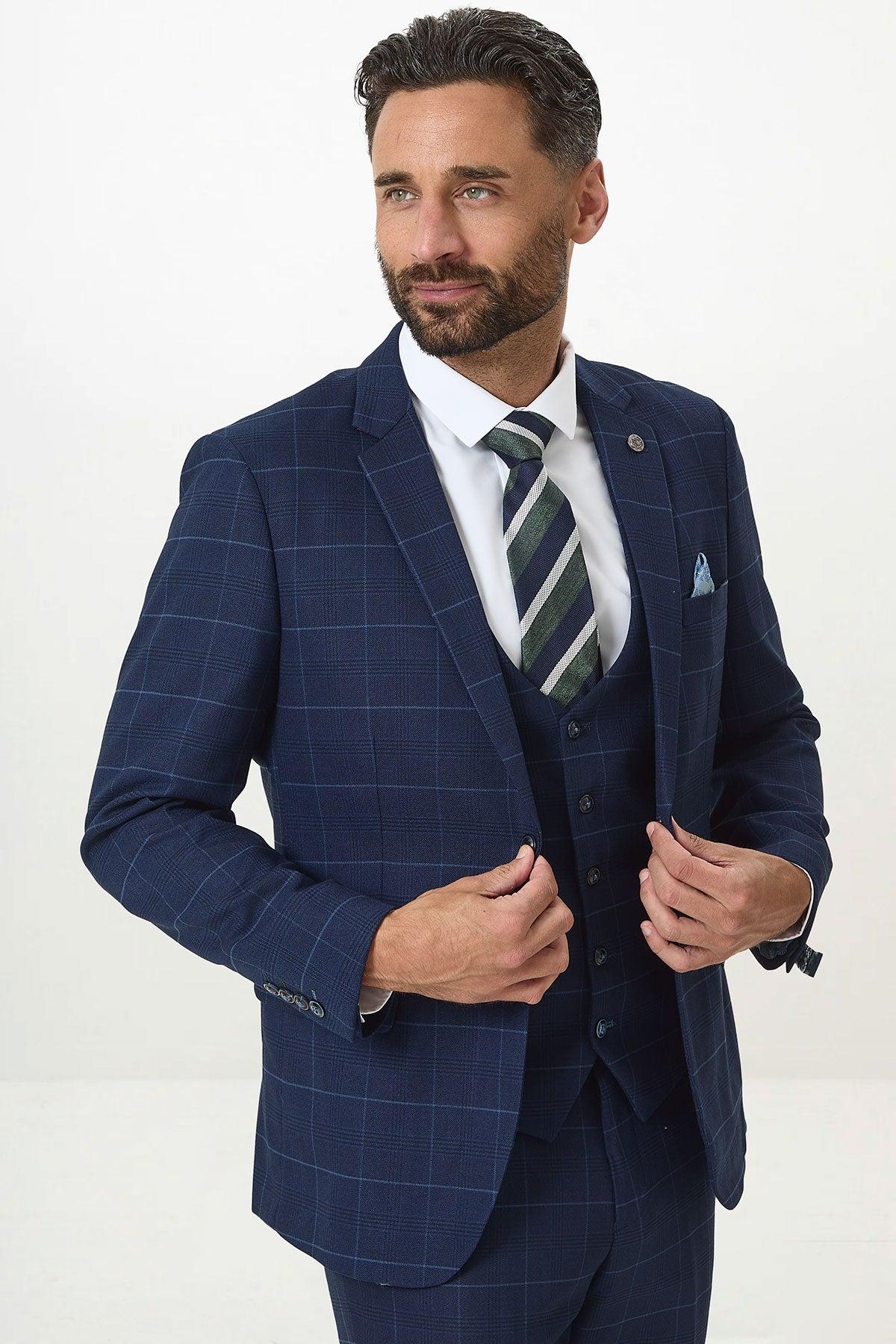 A man with dark hair and a beard wears the Marc Darcy EDINSON Navy Sky Check Three Piece Suit in a slim fit, paired with a white shirt and striped tie, posing with one hand adjusting his jacket against a plain light background.