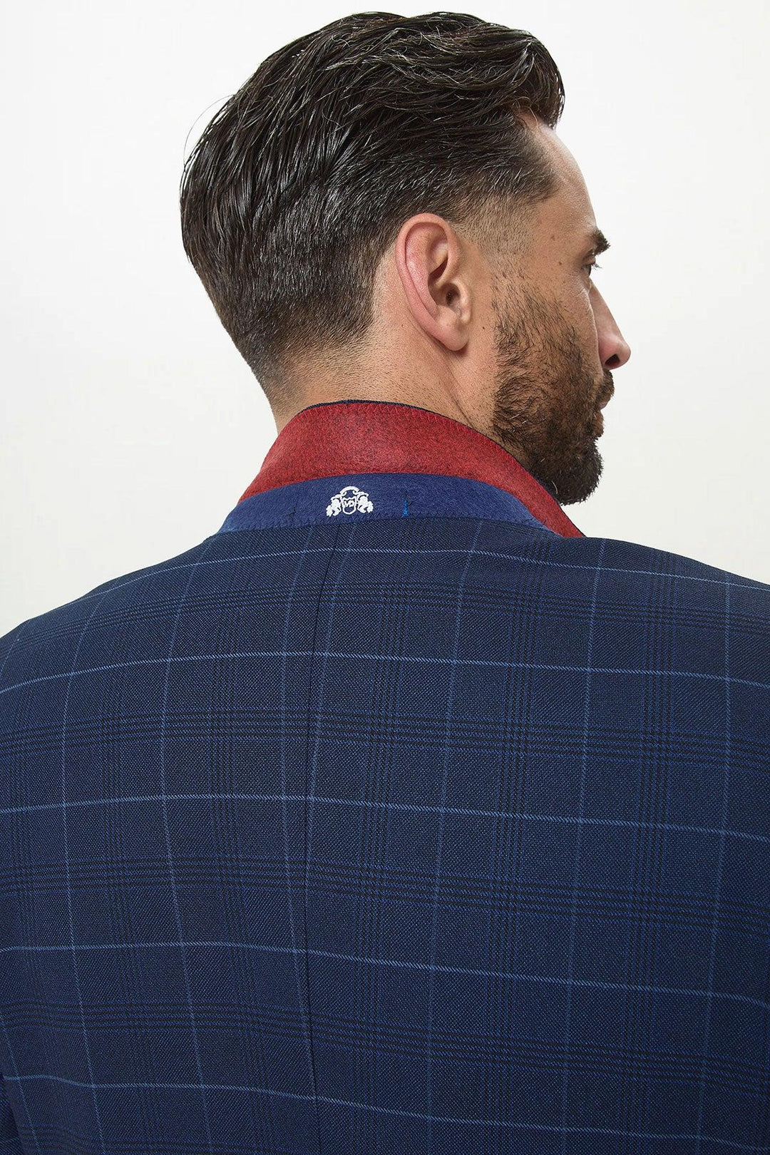 A man with short dark hair and a beard is seen from behind wearing the Marc Darcy EDINSON Navy Sky Check Blazer with a raised collar, featuring a red inner lining and small white embroidery.