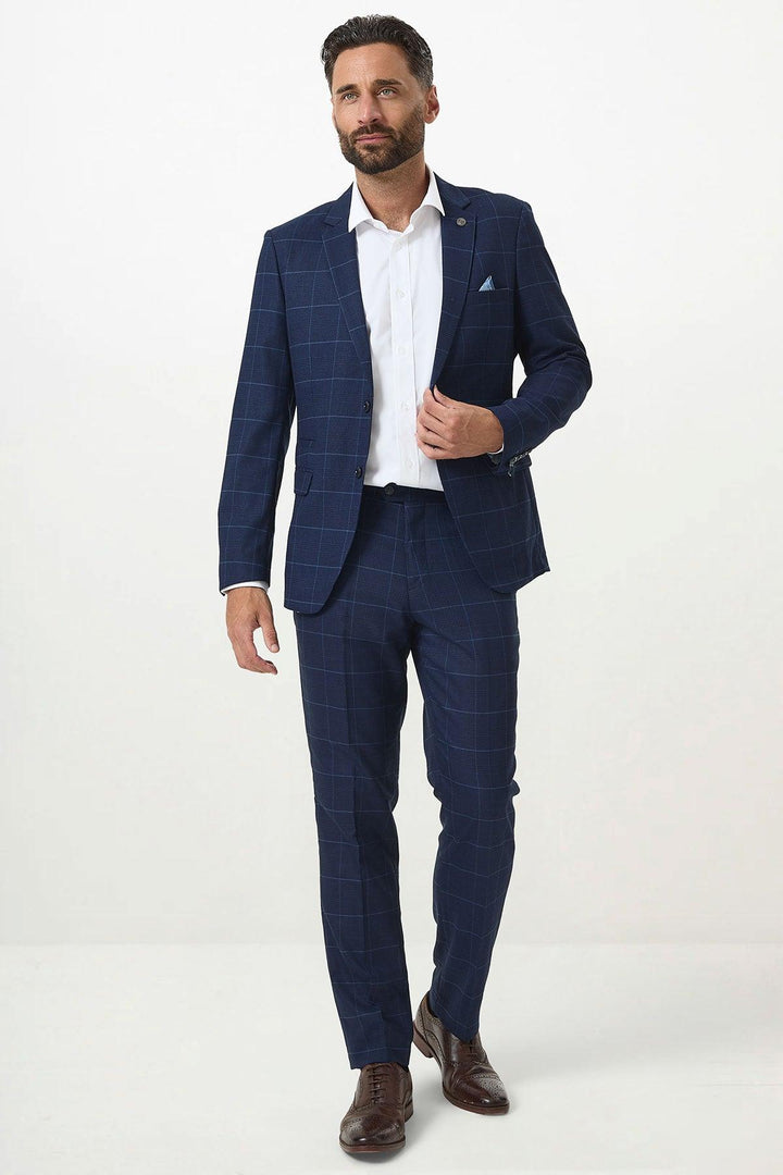 A man models the Marc Darcy EDINSON - Navy Sky Check Blazer over a white dress shirt with brown dress shoes, standing against a plain white background. He adjusts his jacket with one hand and gazes slightly to the side.