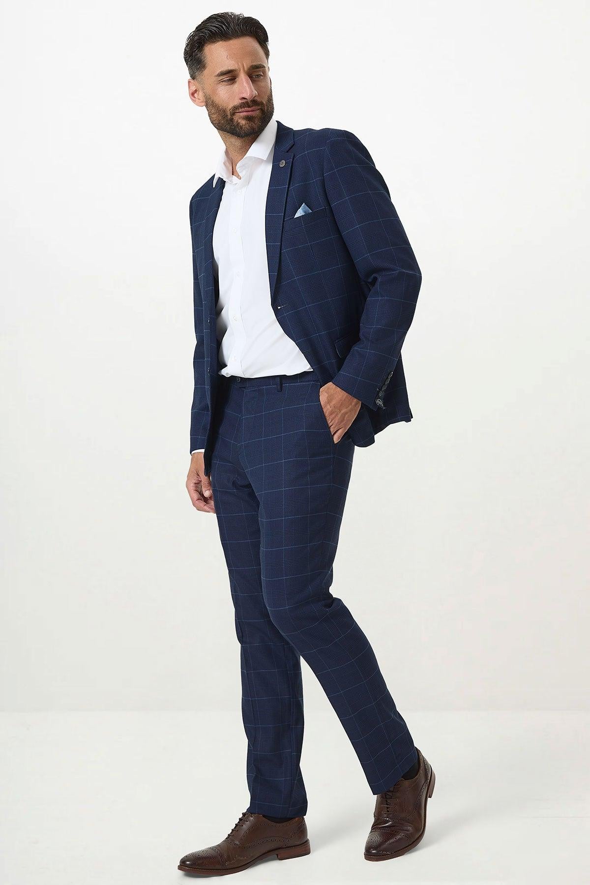 A man stands against a plain white background wearing the Marc Darcy EDINSON Navy Sky Check Two Piece Suit, paired with a white dress shirt, brown dress shoes, and a light blue pocket square.