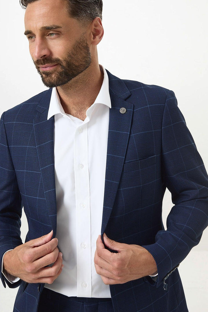 A man with short brown hair and a beard models the Marc Darcy EDINSON Navy Sky Check Blazer over a white dress shirt, holding the lapel and looking to the side against a plain background.