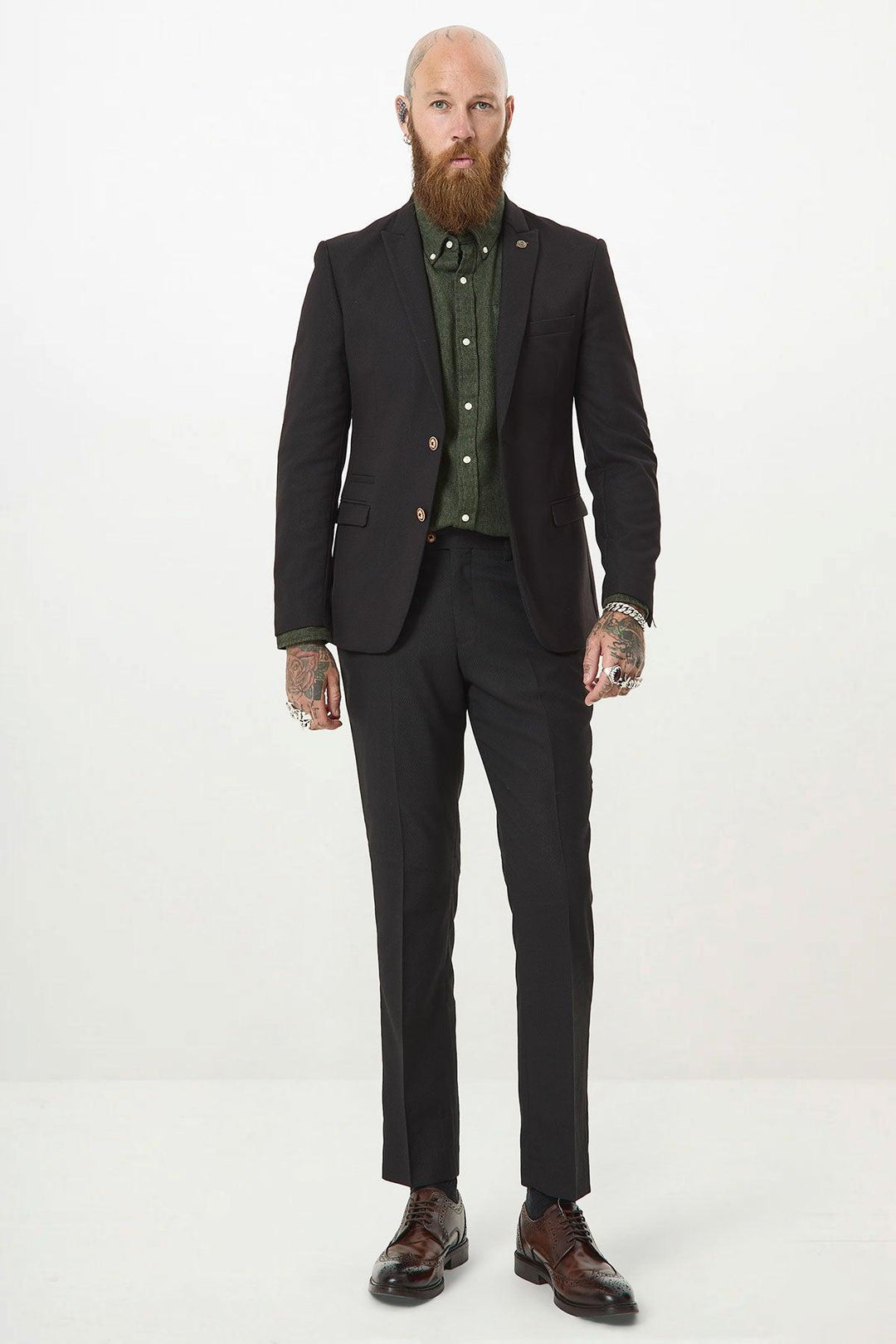 A bearded, tattooed man stands facing forward in a marcdarcy MAX black two piece suit, paired with a green dress shirt and brown dress shoes, against a plain white background.