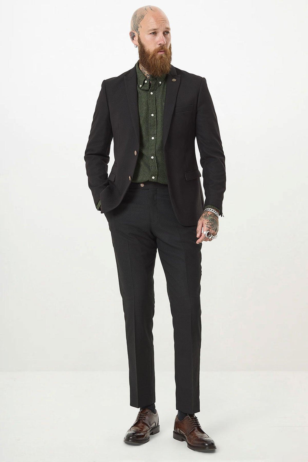 A bearded, tattooed man stands against a white background wearing the marcdarcy MAX Black Two Piece Suit with a green shirt, brown dress shoes, and a camouflage print pocket square for added style. One hand rests in his pocket.