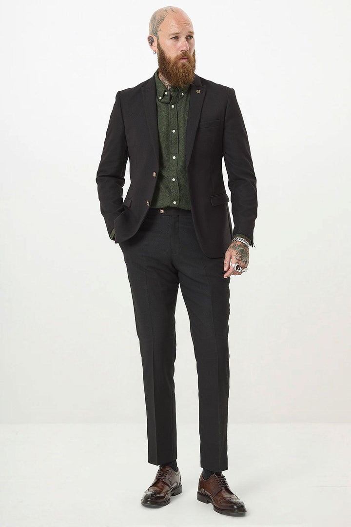 A bearded, tattooed man stands against a white background wearing the marcdarcy MAX Black Two Piece Suit with a green shirt, brown dress shoes, and a camouflage print pocket square for added style. One hand rests in his pocket.