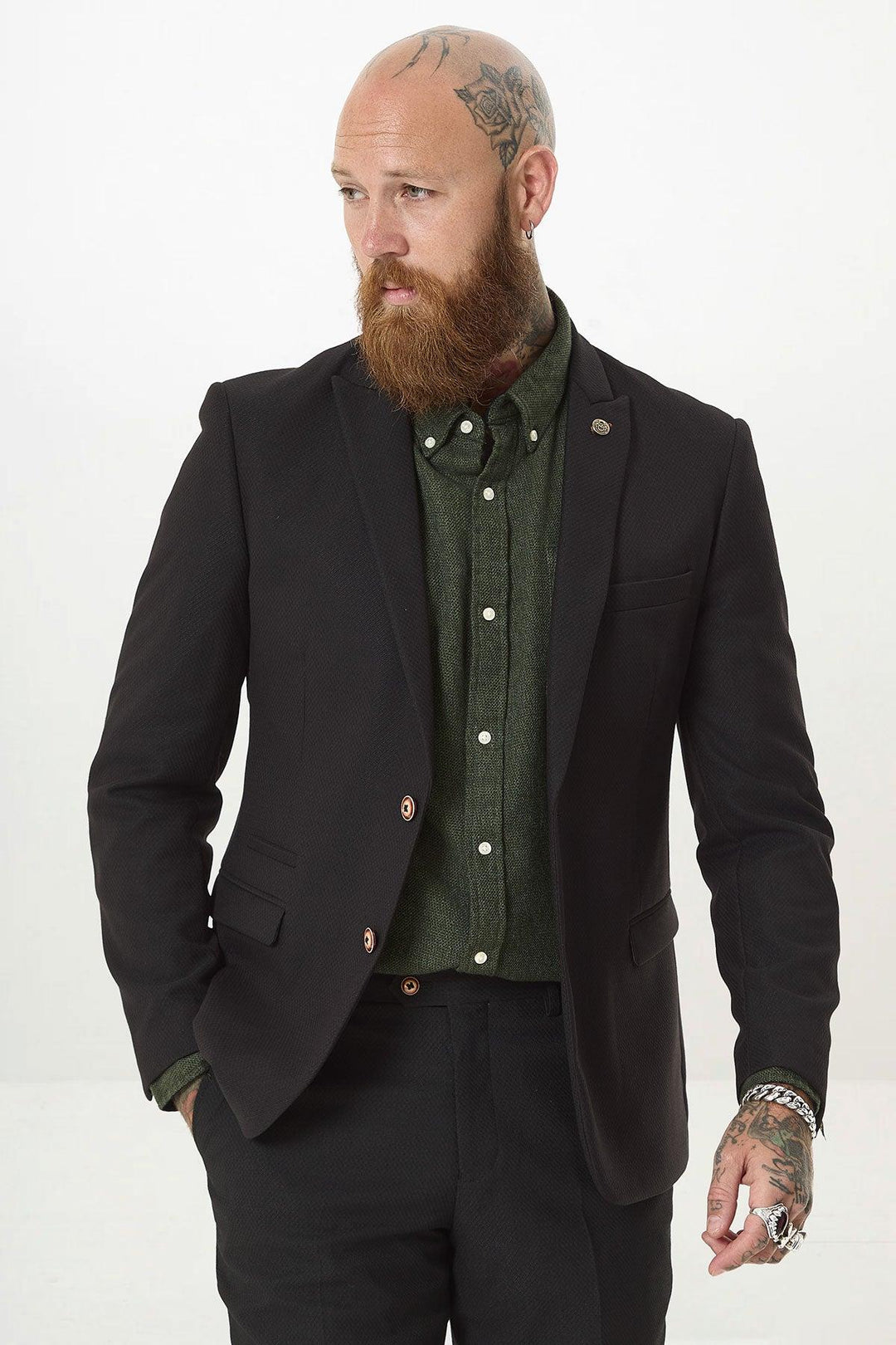 A bearded man with head and hand tattoos wears the marcdarcy MAX Black Two Piece Suit over a green shirt, with a camouflage pocket square. He stands against a white background, looking left with one hand in his pocket.