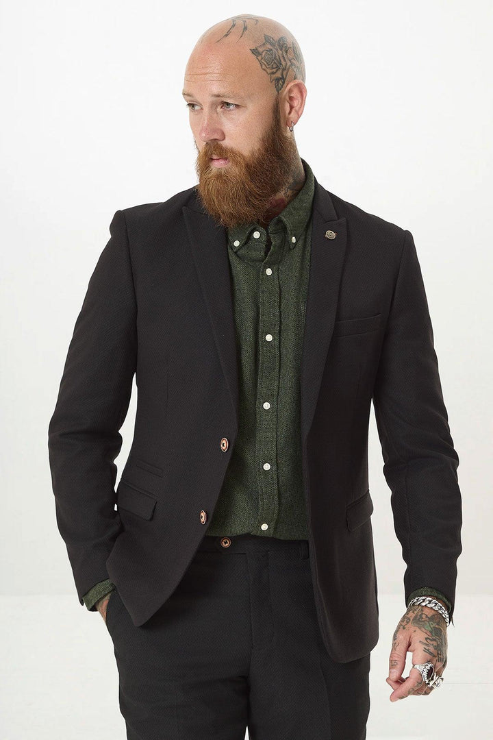A bearded man with head and hand tattoos wears the marcdarcy MAX Black Two Piece Suit over a green shirt, with a camouflage pocket square. He stands against a white background, looking left with one hand in his pocket.
