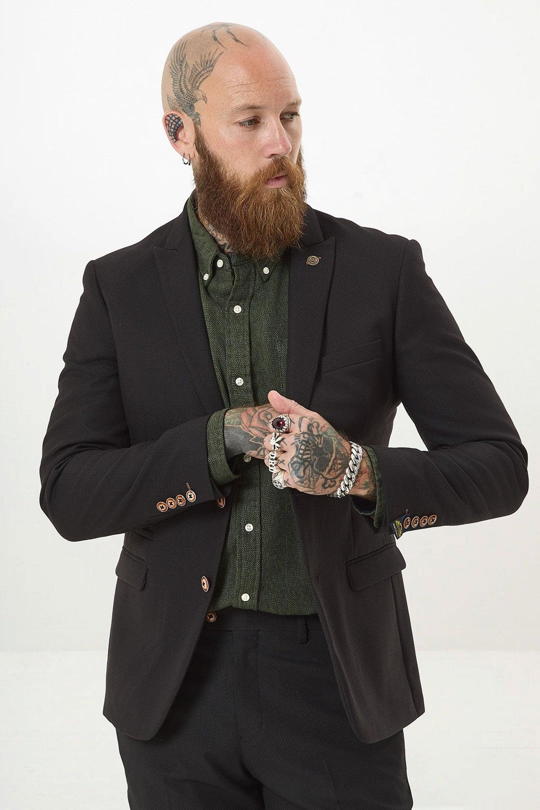A bearded, tattooed, bald man wears the marcdarcy MAX black two piece suit over a dark green shirt with a camouflage pocket square, accessorized with rings and bracelets, looking sideways against a plain white background.