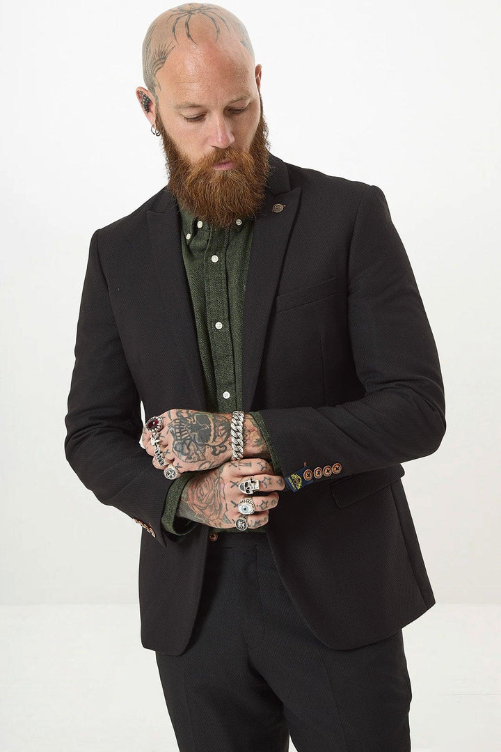 A bald man with a red beard and tattoos wears the marcdarcy MAX Black Two Piece Suit over a green shirt. He looks down, adjusting his sleeve to reveal rings and bracelets. The background is plain white.