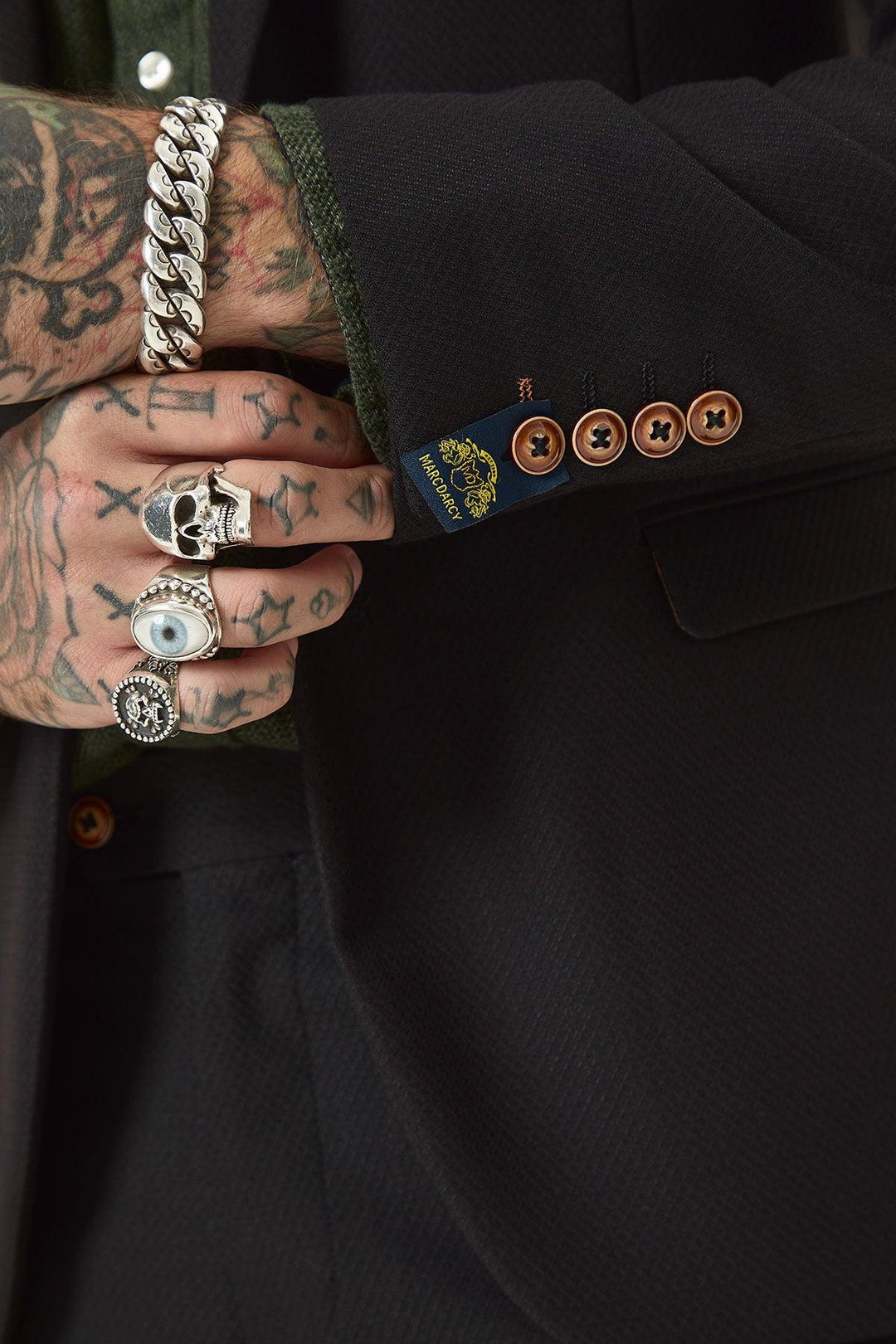 A tattooed hand with rings holds the cuff of a marcdarcy MAX Black Two Piece Suit blazer, featuring brown buttons and a visible designer label, paired with a green shirt.