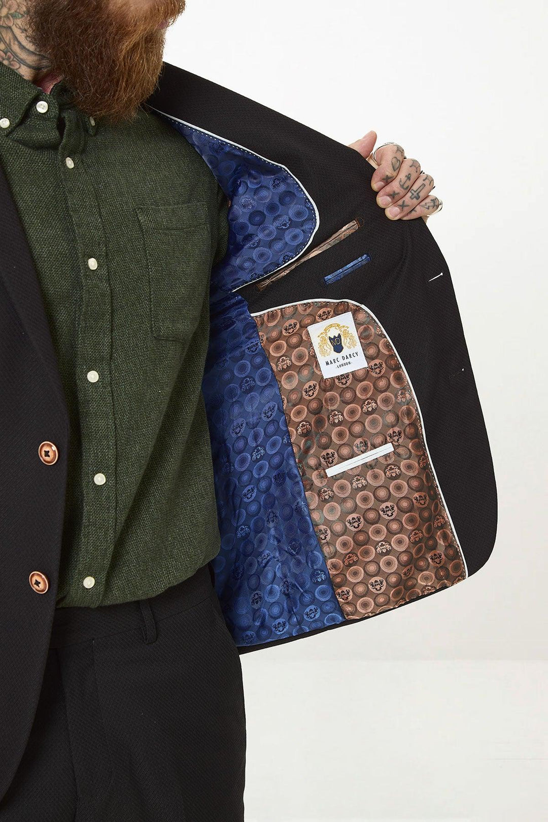 A bearded, tattooed man opens his marcdarcy MAX - Black Two Piece Suit blazer to show a skull-patterned lining and labels. He pairs it with a green shirt, black pants, and a camouflage pocket square for extra style.