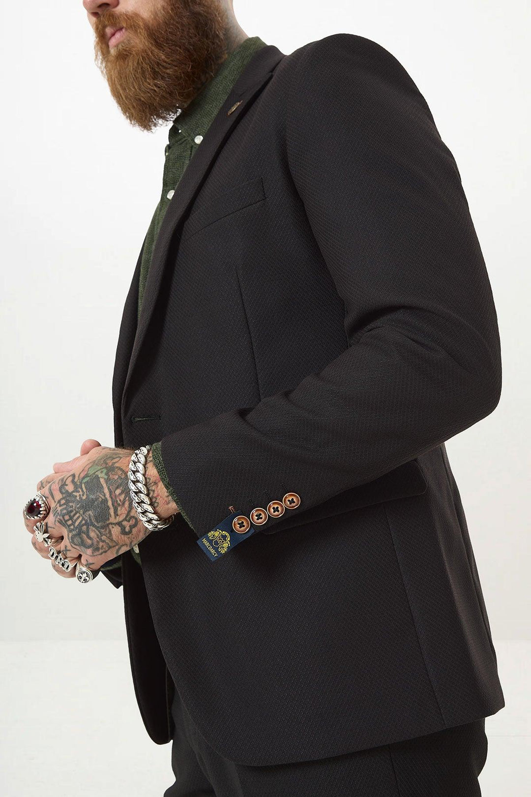 A man with a beard and tattooed hand, wearing rings and bracelets, models the marcdarcy MAX Black Two Piece Suit—slim fit blazer over a green shirt with contrasting brown buttons and a camouflage pocket square. Upper body only, plain background.