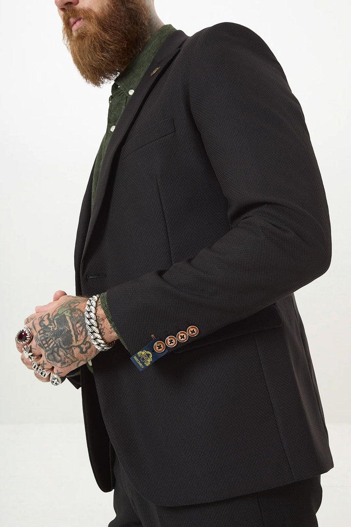 A man with a beard and tattooed hand, wearing rings and bracelets, models the marcdarcy MAX Black Two Piece Suit—slim fit blazer over a green shirt with contrasting brown buttons and a camouflage pocket square. Upper body only, plain background.