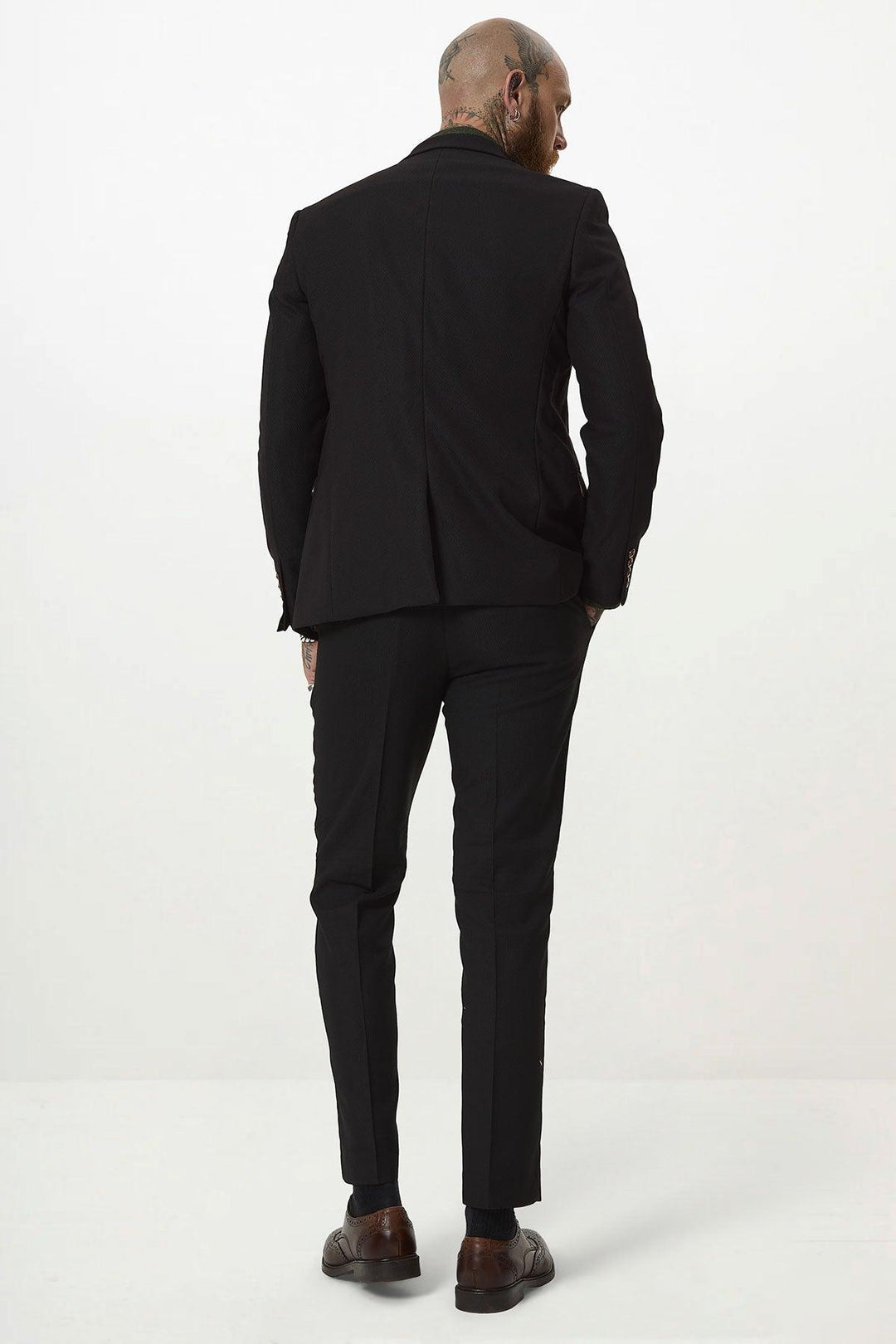 A bald, tattooed man stands facing away against a plain white background, wearing the marcdarcy MAX Black Two Piece Suit with brown shoes and a camouflage print pocket square for added flair.