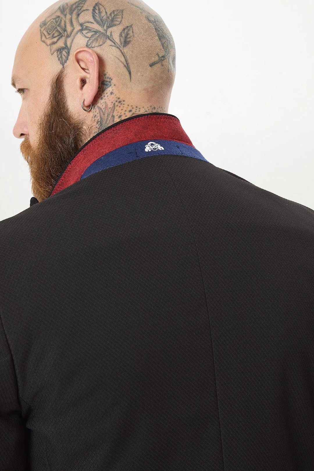 A bald man with a floral scalp tattoo and ear piercing is seen from the back and side wearing the marcdarcy MAX Black Two Piece Suit, featuring a slim fit, red and blue collar detail, and a camouflage print pocket square.