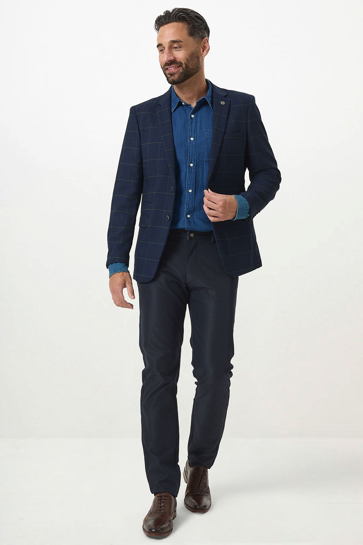 A man in a Marc Darcy EDINSON Navy Green Check Blazer, blue denim shirt, and navy pants smiles and adjusts his blazer against a white background.