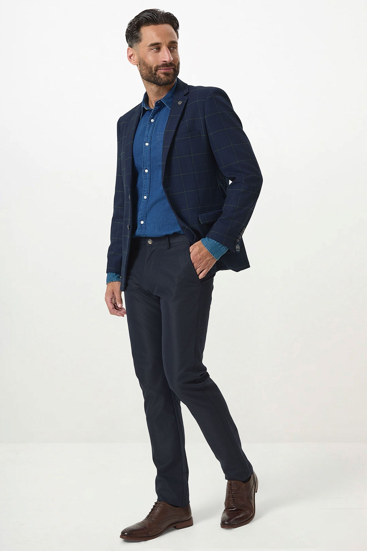 A man with short dark hair and a beard stands against a white background, wearing the Marc Darcy EDINSON Navy Green Check Blazer over a blue shirt, paired with dark trousers and brown dress shoes. He looks to the side with a slight smile.