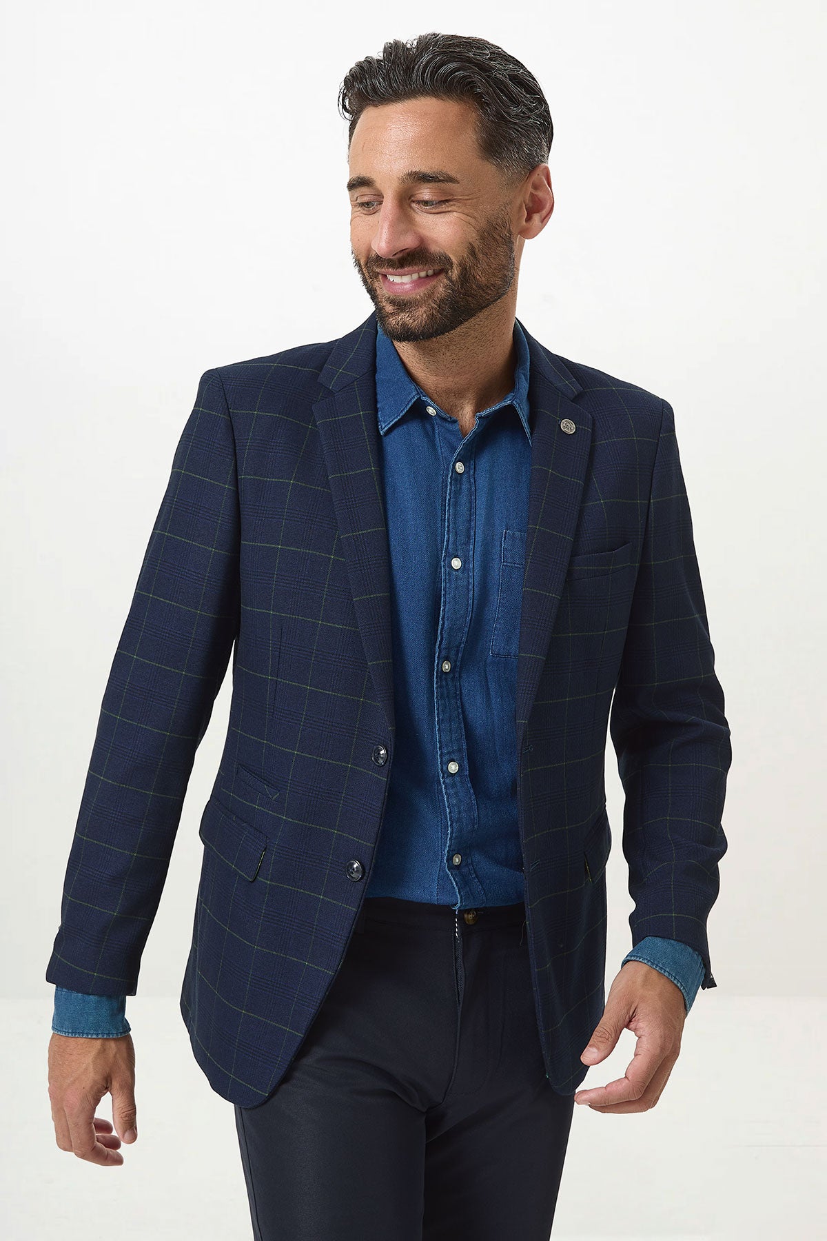 A man with dark hair and a beard wears the Marc Darcy EDINSON Navy Green Check Blazer over a blue denim shirt and dark trousers. He smiles and looks to the side against a plain white background.