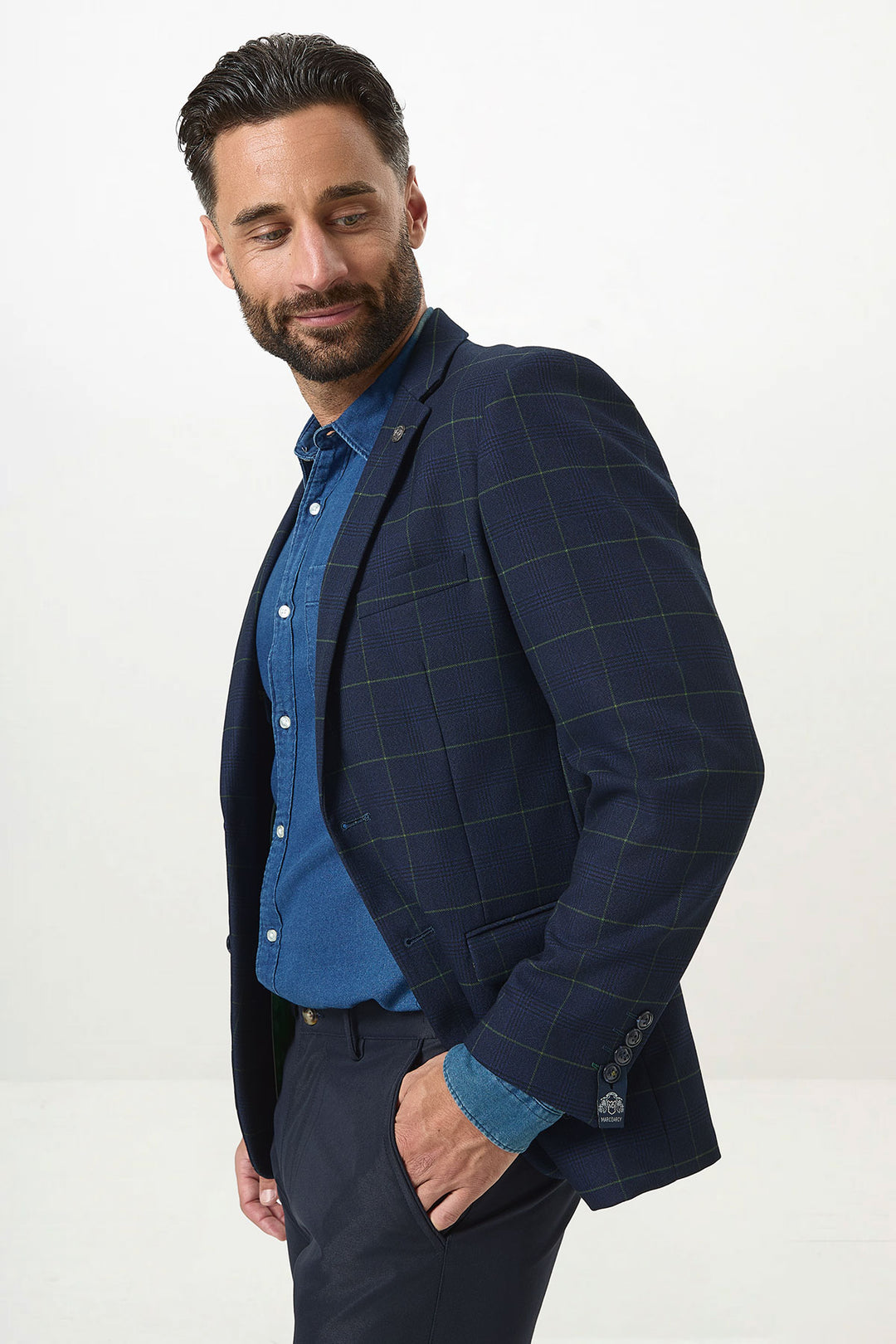 A man with dark hair and a beard wears the Marc Darcy EDINSON Navy Green Check Blazer over a blue shirt and dark pants, standing with one hand in his pocket and glancing over his shoulder, smiling slightly against a plain background.