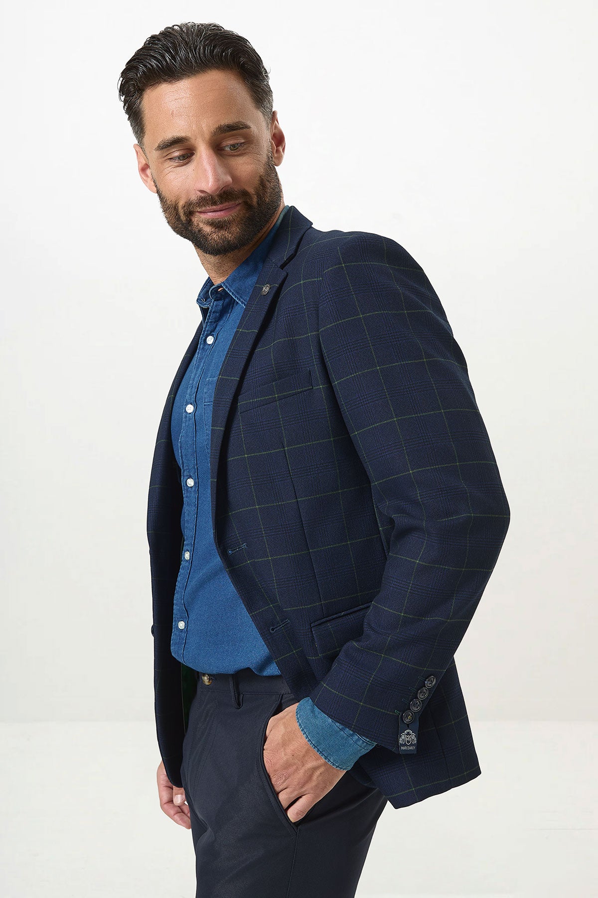 A man with a beard wears the Marc Darcy EDINSON Navy Green Check Blazer over a denim shirt and navy trousers, his hand casually in his pocket.