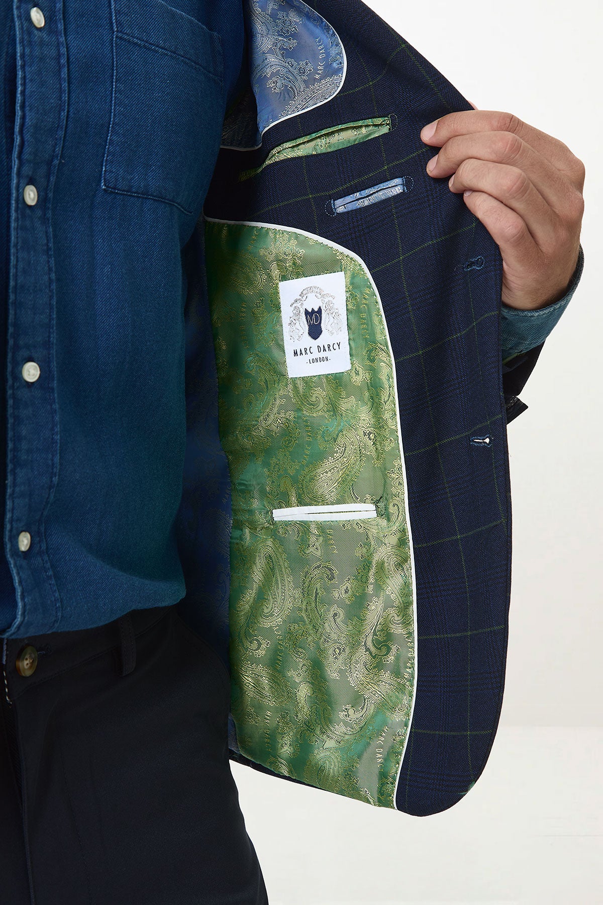 A man in a blue denim shirt and black pants displays the inside of the Marc Darcy EDINSON Navy Green Check Blazer, featuring a green paisley lining and a Marc Darcy label.