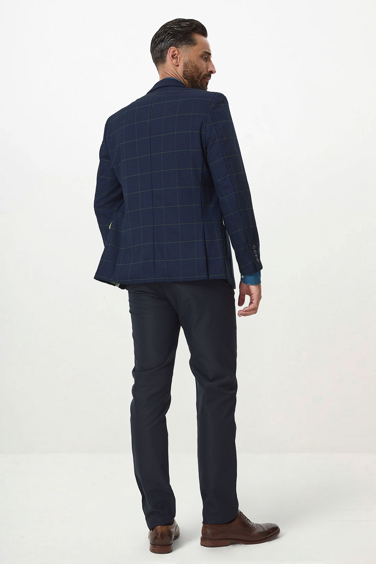 A man with dark hair and a beard wears the Marc Darcy EDINSON Navy Green Check Blazer, paired with dark trousers, brown shoes, and a light blue shirt. He stands against a plain white background, facing away while looking over his shoulder.