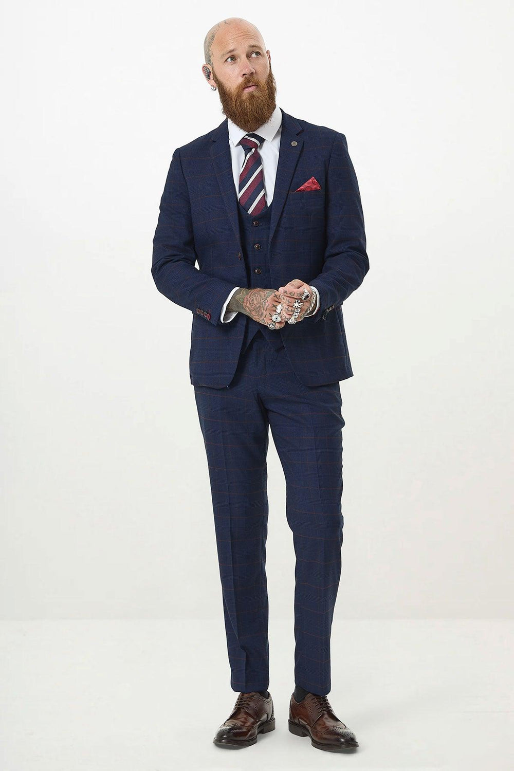 A bearded man in the Marc Darcy EDINSON Navy Wine Check Three Piece Suit stands against a white background, wearing a red striped tie and pocket square, gazing upward with his tattooed hands clasped confidently.