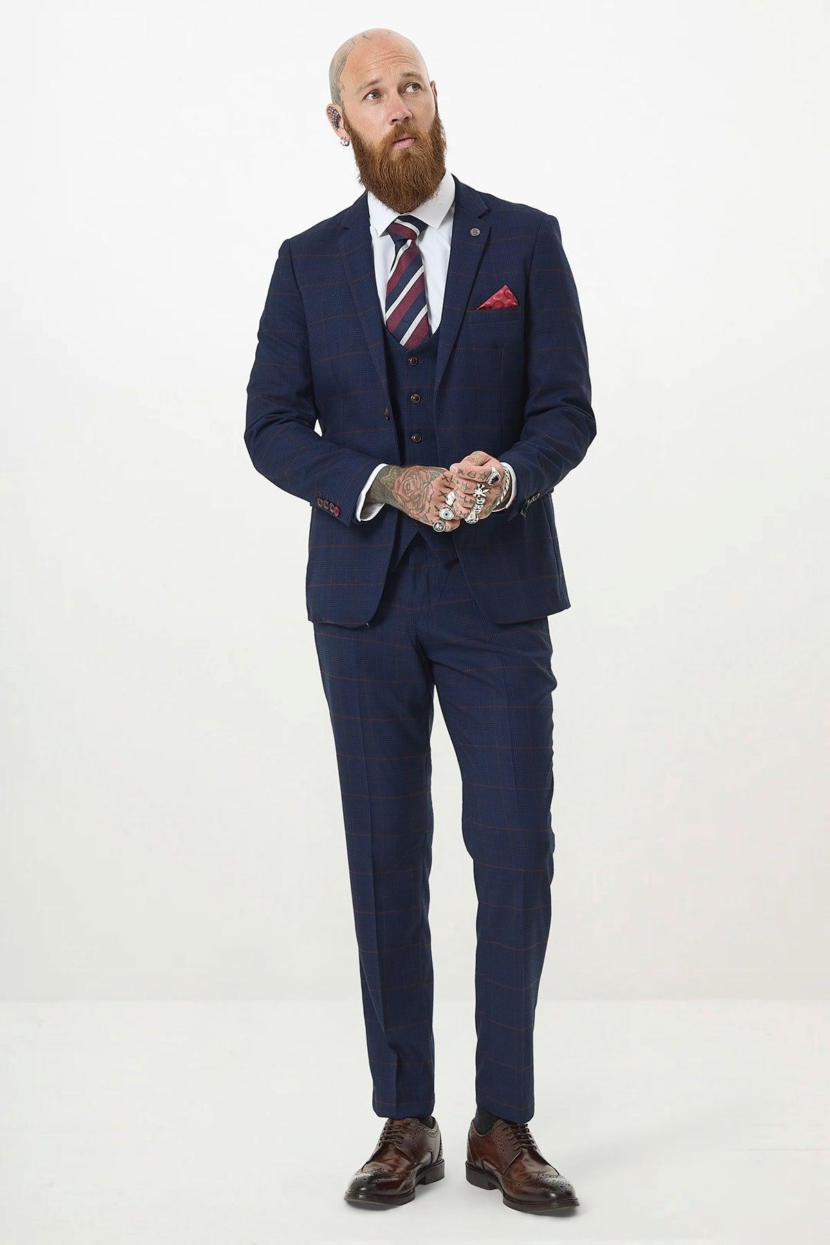 A bearded man with hand tattoos wears the Marc Darcy EDINSON Navy Wine Check Three Piece Suit with a white shirt, striped tie, and brown dress shoes. He stands against a plain white background, looking upward with hands clasped.