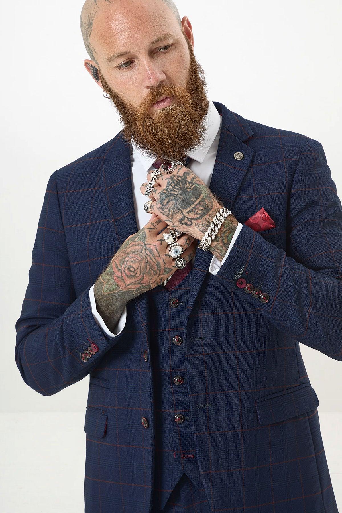 A man wearing the Marc Darcy EDINSON Navy Wine Check Three Piece Suit adjusts his tie, showing tattoos on his arms and hands, and accessorizes with multiple rings and a bracelet.