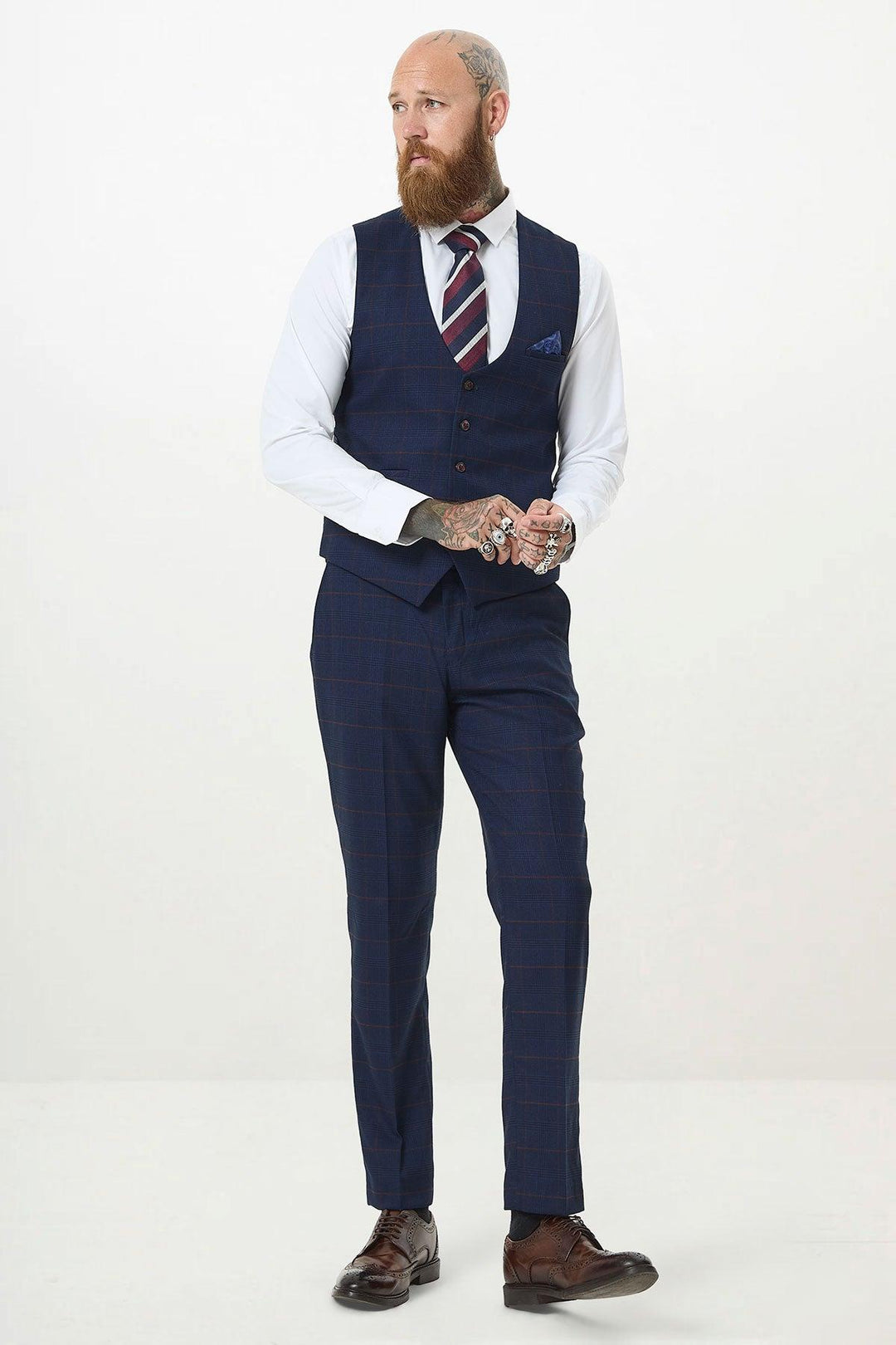 A bearded, tattooed man in a white shirt, Marc Darcy EDINSON navy wine check single breasted waistcoat, matching trousers, striped tie, and brown dress shoes stands against a plain white background.
