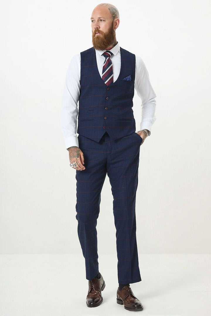 A bearded, tattooed man wears a Marc Darcy EDINSON navy wine check single breasted waistcoat with a white shirt, striped tie, brown shoes, and slim fit trousers, standing against a plain white background.
