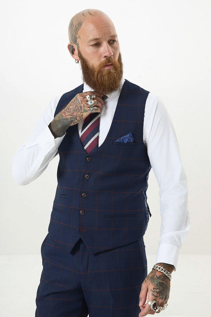 A bearded, tattooed man with a bald head adjusts his Marc Darcy EDINSON Navy Wine Check Single Breasted Waistcoat, paired with a striped tie, white shirt, and matching plaid pants. He wears rings, bracelets, an earring, and looks slightly to the side.