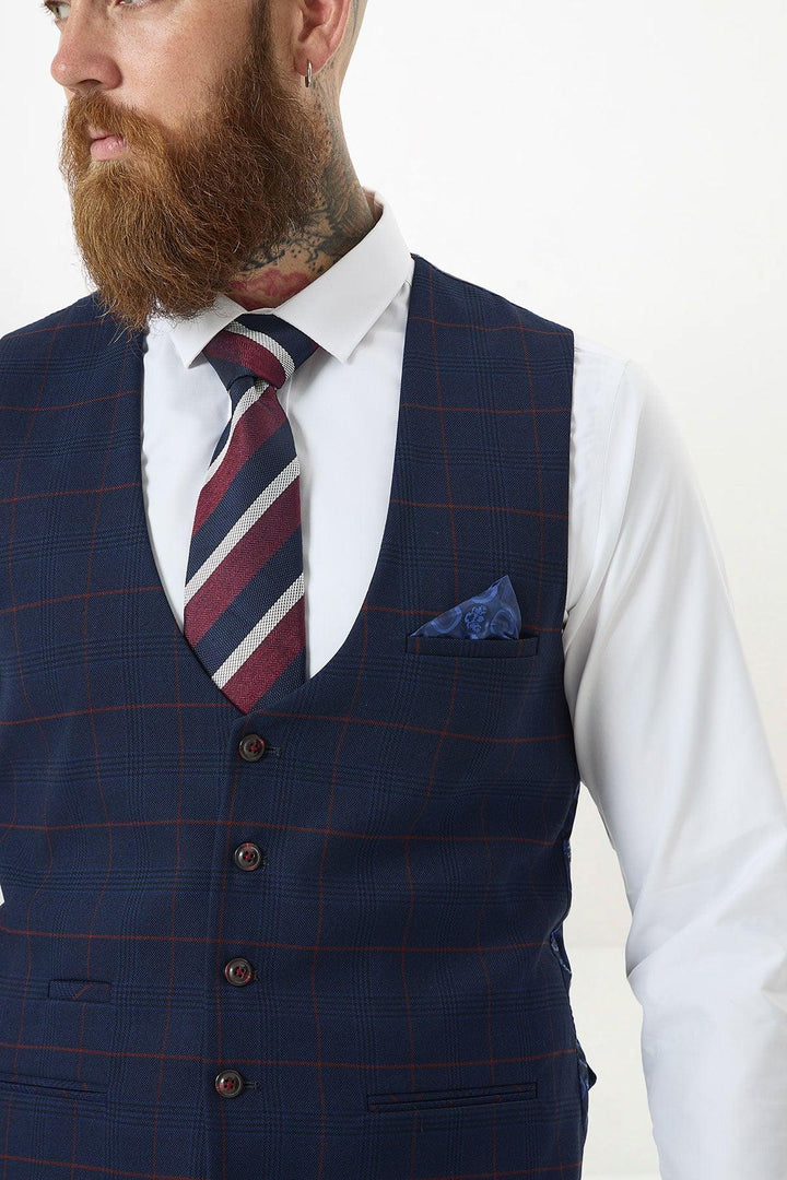 A bearded man with neck tattoos wears the Marc Darcy EDINSON Navy Wine Check single-breasted waistcoat over a white shirt and blue/red striped tie, accented by a blue pocket square, against a plain light background.
