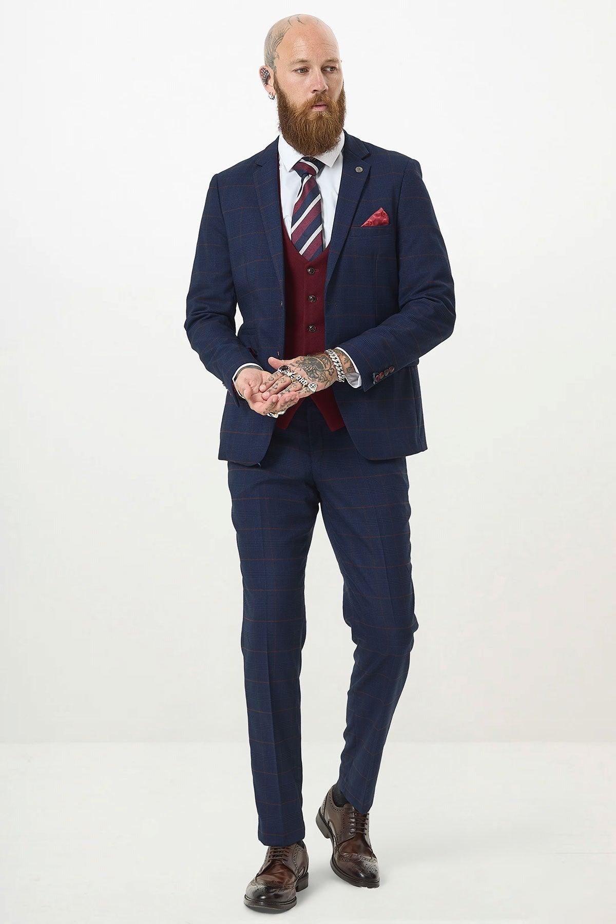 A bearded man with hand tattoos wears the Marc Darcy EDINSON Navy Check Suit with a Kelvin Wine Waistcoat, white shirt, striped tie, and brown dress shoes. He stands against a plain white background.