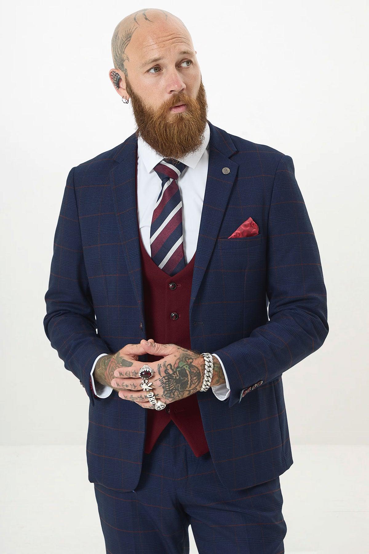 A bearded man with tattoos and piercings wears the Marc Darcy EDINSON Navy Check Suit with a Kelvin Wine Waistcoat. He pairs it with a striped tie and pocket square, standing against a plain white background, looking to his right.