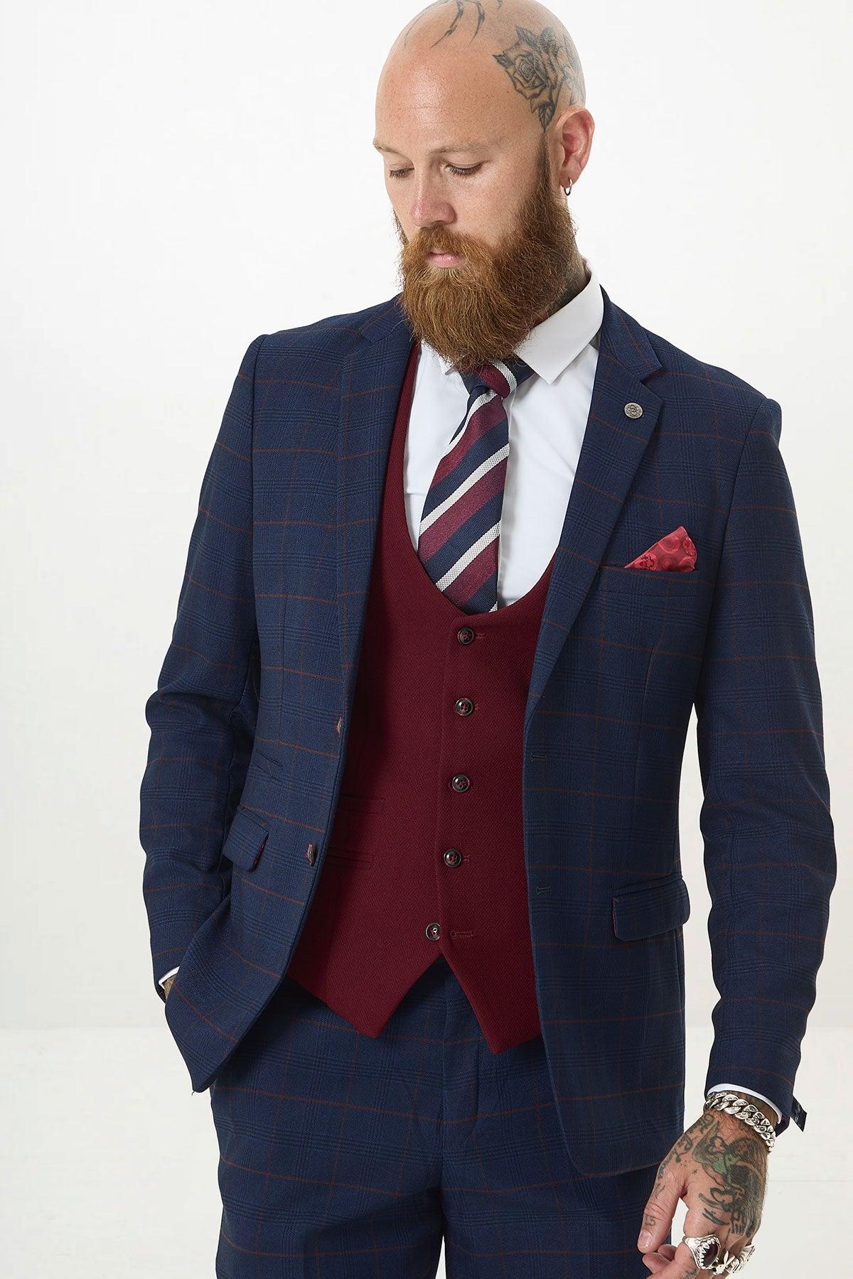 A bearded man with tattoos and piercings wears Marc Darcy’s EDINSON Navy Check Suit with a Kelvin Wine Waistcoat, white shirt, and striped tie. He looks down with one hand in his pocket against a plain white background.
