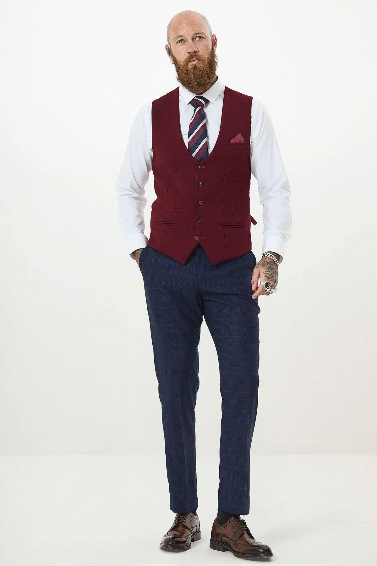 A bearded man with tattoos stands against a white background wearing the marcdarcy KELVIN Wine Single Breasted Waistcoat, a white shirt, striped tie, blue checked trousers, brown dress shoes, and a pocket square in his slim fit waistcoat.