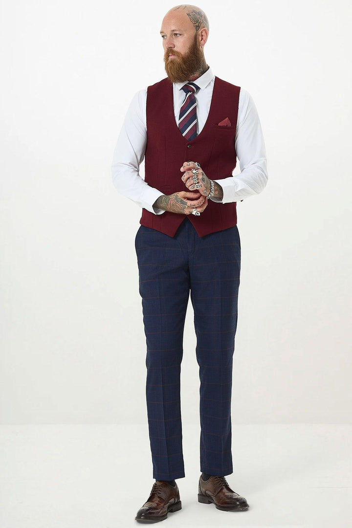 A bearded man with tattoos wears the marcdarcy KELVIN - Wine Single Breasted Waistcoat over a white shirt, striped tie, navy checkered pants, and brown dress shoes, standing against a plain white background.