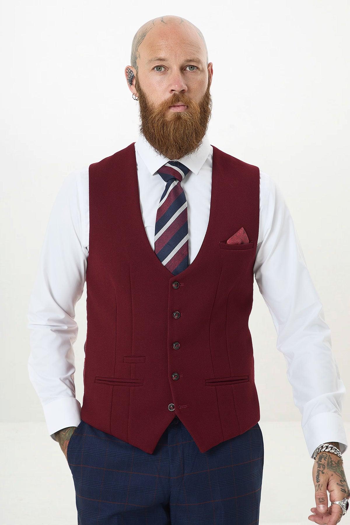 A bald, tattooed man with a bushy red beard models the marcdarcy KELVIN - Wine Single Breasted Waistcoat over a white shirt, striped tie, and dark plaid trousers, accented with a pocket square against a plain white background.