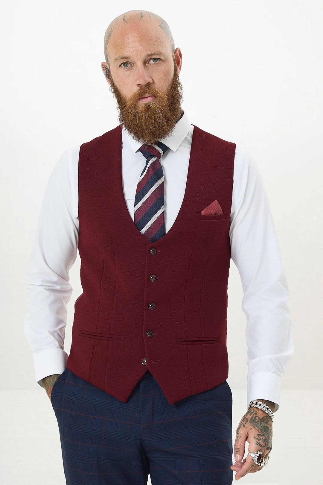 A bearded, tattooed man stands with hands in pockets, wearing marcdarcys KELVIN - Wine Single Breasted Waistcoat over a white shirt, striped tie, pocket square, and navy plaid pants against a plain white background.