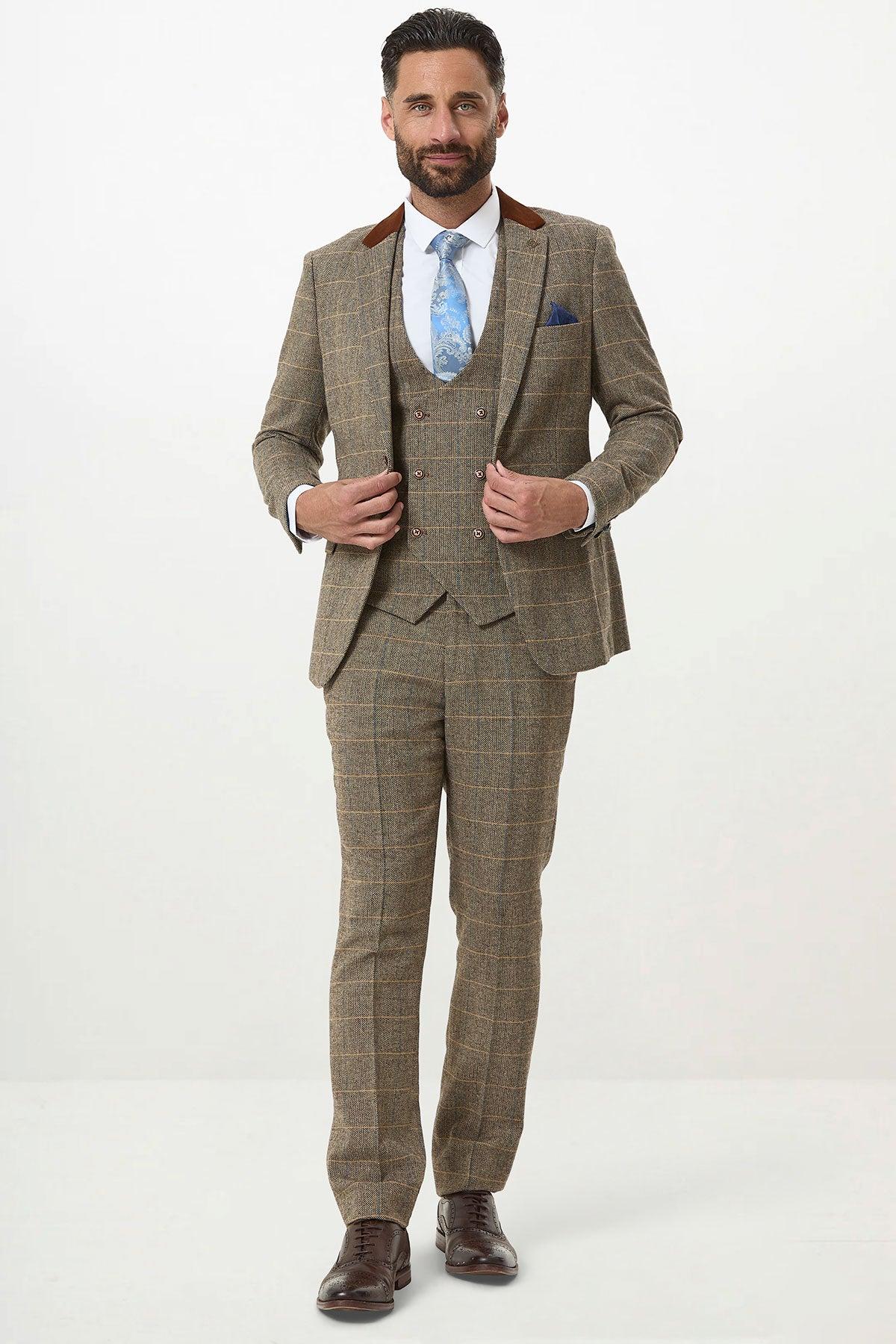 A man stands facing forward, smiling slightly in the marcdarcy DX7 Tan Tweed Check Three Piece Suit with double-breasted waistcoat, paired with a blue tie, white shirt, brown shoes, and a blue pocket square. He holds his jacket with both hands.