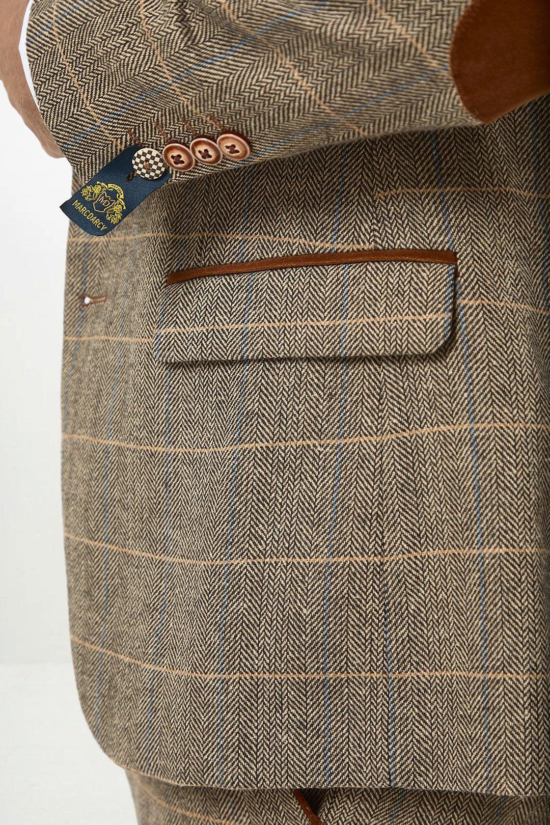 Close-up of a person wearing the marcdarcy DX7 Tan Tweed Check Suit with Kelvin Cream Waistcoat, featuring brown trim pockets, elbow patches, and four brown buttons with a patterned blue tag on the sleeve cuff—perfect for rustic country weddings.
