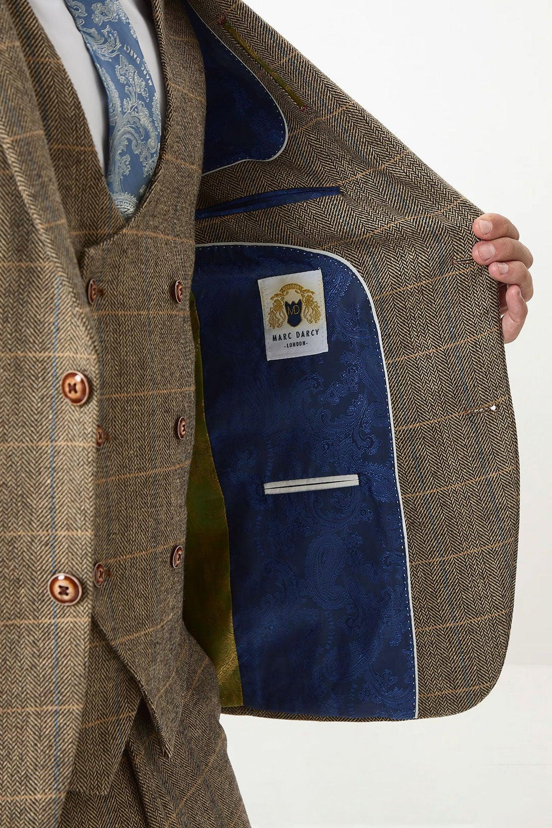 A person in a tan tweed check suit with a Kelvin cream waistcoat by marcdarcy holds open the jacket to show a blue paisley lining and label, teamed with a blue patterned tie—perfect for a rustic country wedding look.