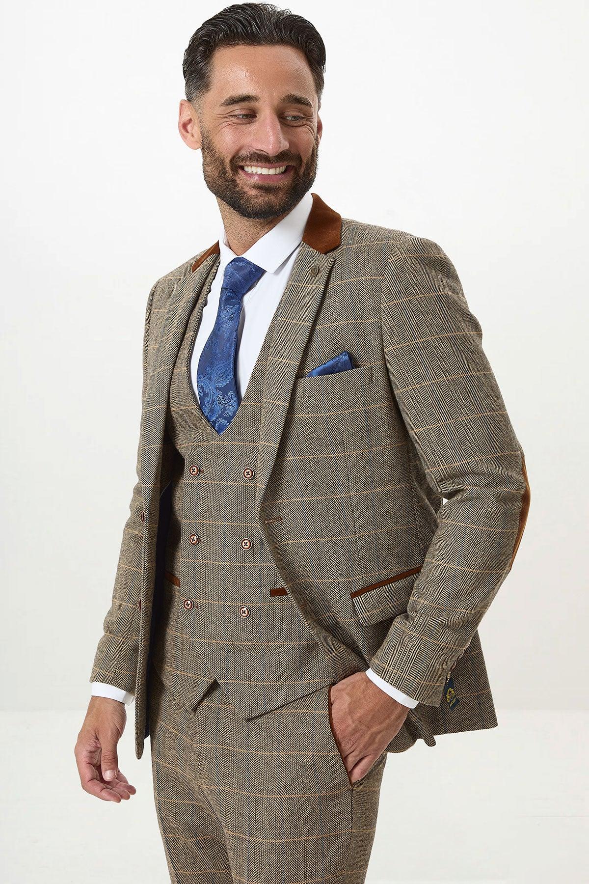 A man with dark hair and a beard smiles in the marcdarcy DX7 Tan Tweed Check Three Piece Suit with double-breasted waistcoat, white shirt, and blue patterned tie, his left hand in his pocket against a plain white background.