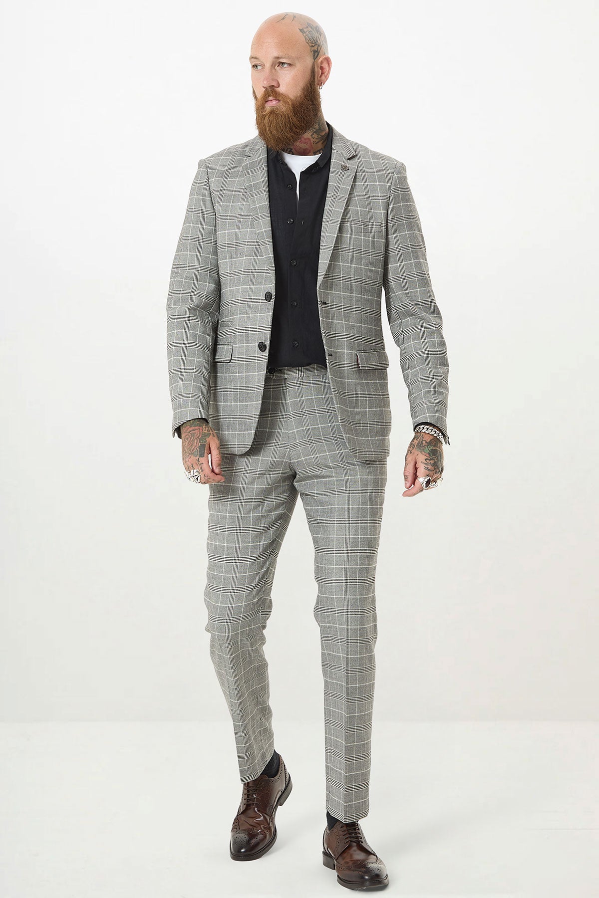 A man with a beard and tattoos wears the Marc Darcy ROSS Grey Check Two Piece Suit, paired with a black shirt and brown dress shoes, standing against a plain white background.