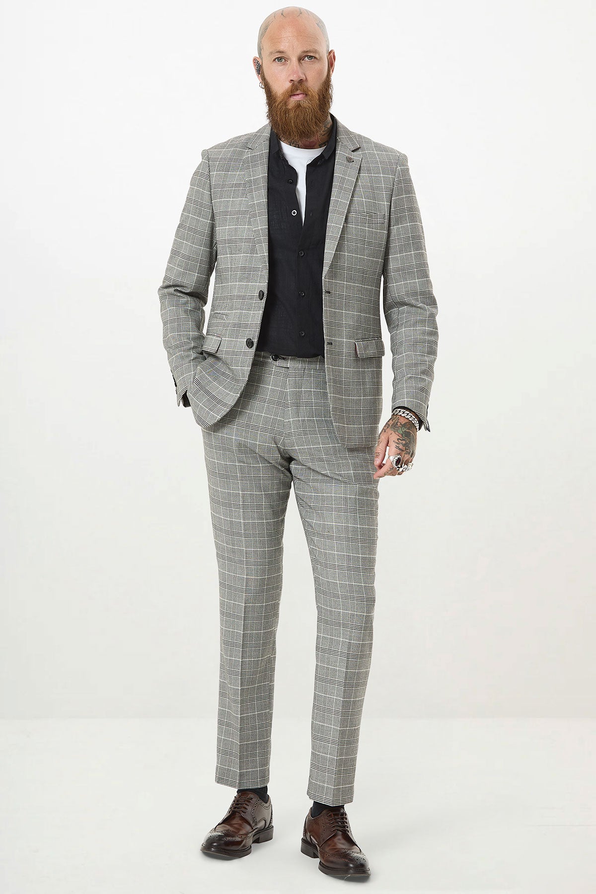 A tattooed, bald man with a beard wears the Marc Darcy ROSS Grey Check Two Piece Suit, a black shirt, and brown dress shoes.