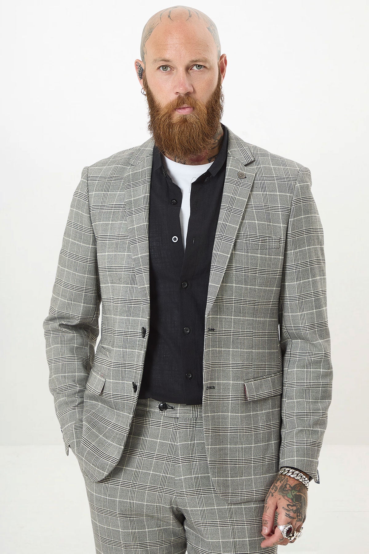 A bearded, tattooed man wears the Marc Darcy ROSS Grey Check Two Piece Suit over a black shirt. He stands against a plain white background with one hand in his pocket, looking directly at the camera.