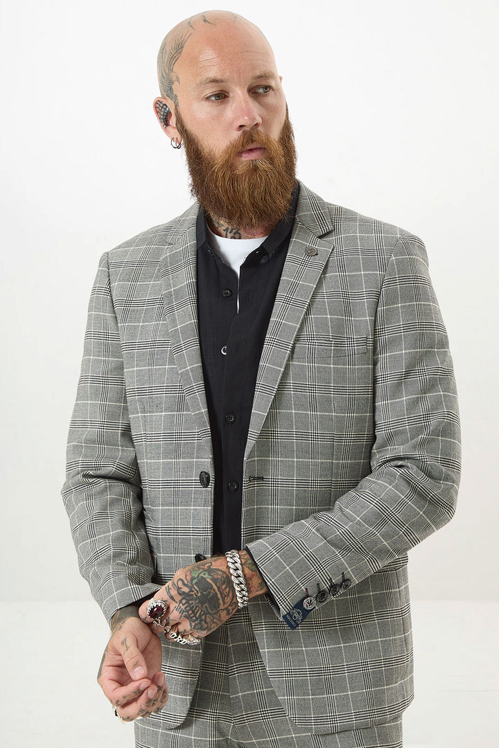 A bearded, tattooed man wears the Marc Darcy ROSS - Grey Check Blazer over a black shirt, standing against a plain white background and looking to the side with his hands together.