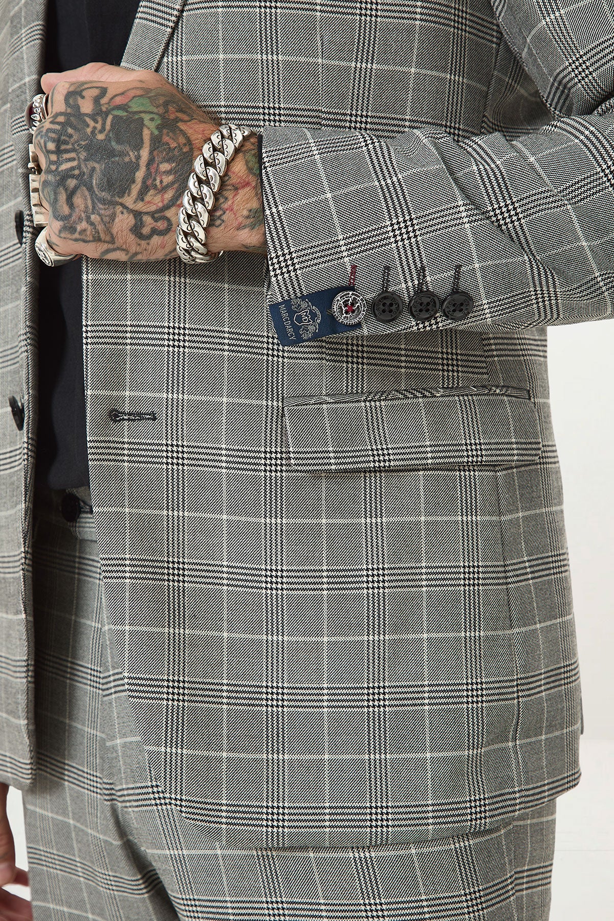 A person wearing the Marc Darcy ROSS Grey Check Two Piece Suit with visible tattoos and chunky silver bracelets stands with one hand near their jacket. The slim-fit suit features four sleeve buttons and a stylish lapel pin.