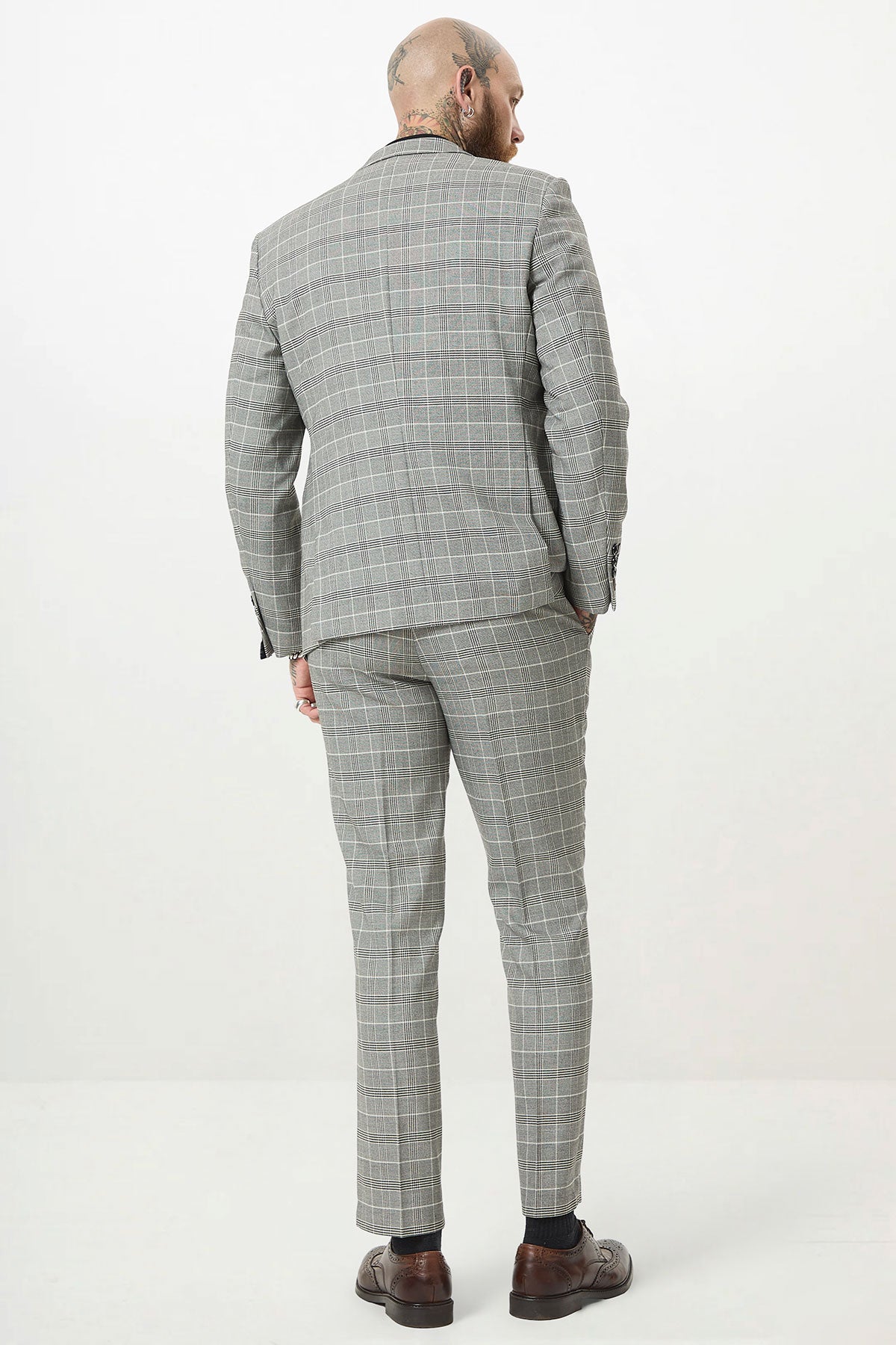 A man with head and neck tattoos stands with his back to the camera, wearing the Marc Darcy ROSS Grey Check Two Piece Suit and brown dress shoes against a plain white background.