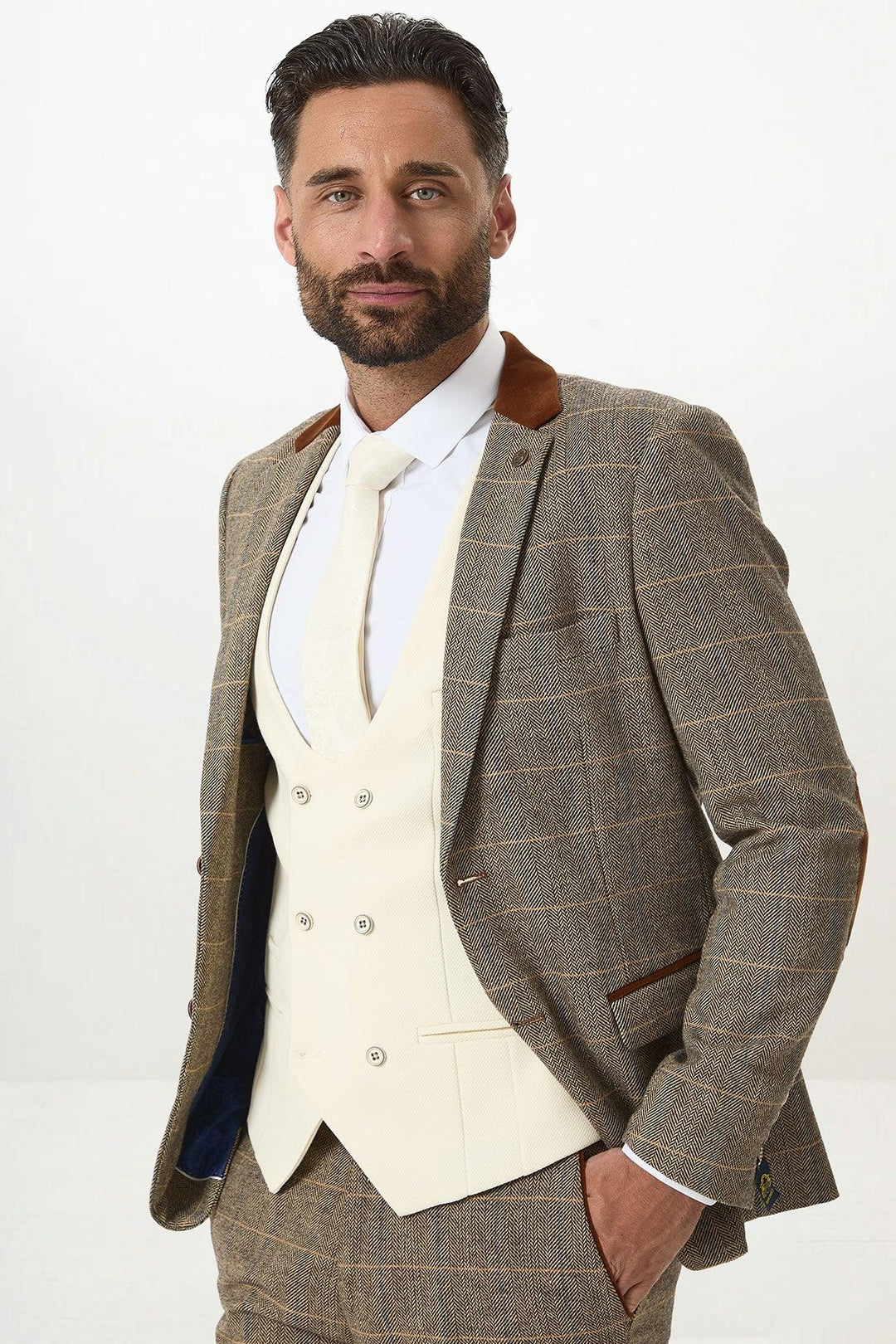A man with dark hair and a beard wears the marcdarcy DX7 Tan Tweed Check Suit with a Kelvin Cream Waistcoat, white shirt, and tie, standing against a plain light background with his hands in his pockets.